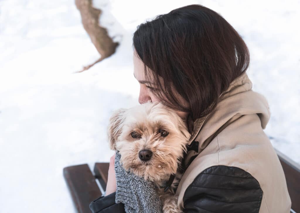 Young Woman Sitting On A Bench Holding Her Havanese Dog With Affection, To Protect It From Cold