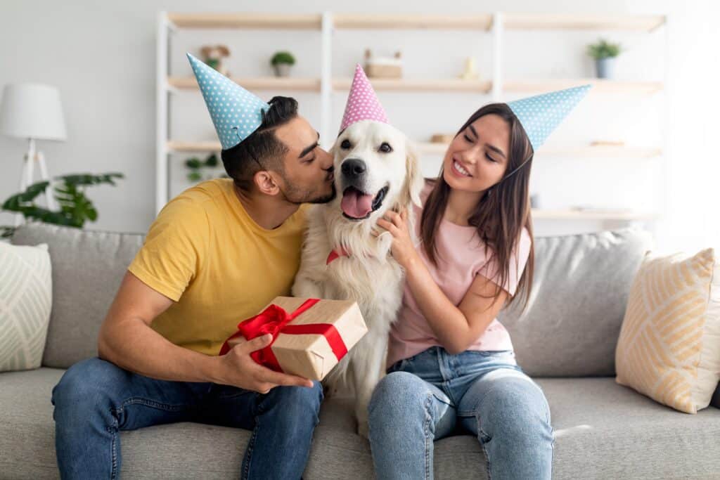 Couple Wearing Party Hats Kissing Their Dog While Holding A Gift